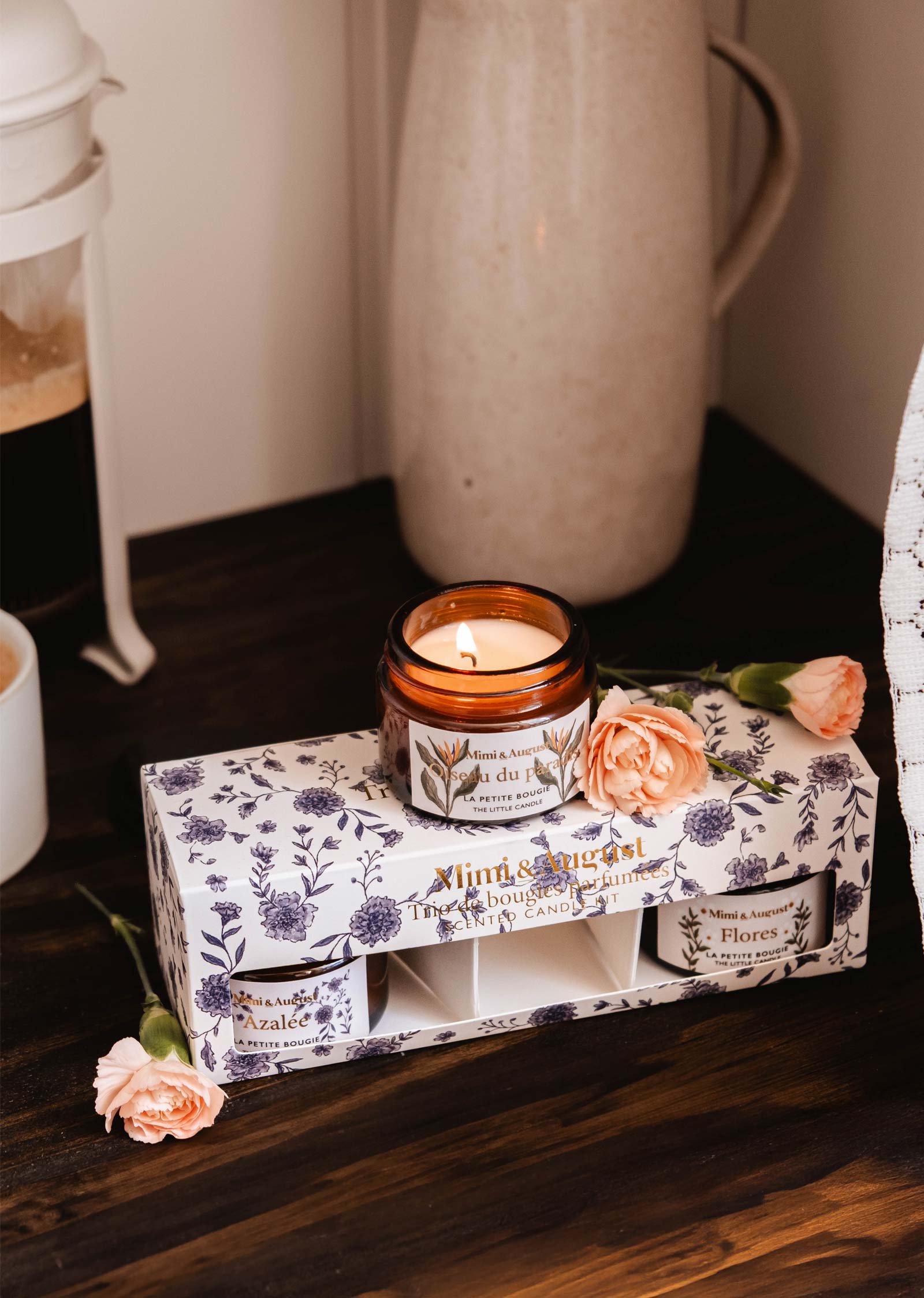 A lit floral candle in a brown jar is placed on The Secret Garden Box Set by Mimi & August, alongside three other candles and two pink carnations, all arranged on a dark wooden table.