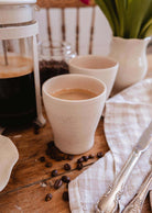 The Small Ceramic Cup by Mimi & August sits on a wooden table with coffee beans, a French press, a napkin, knife, and vase in the background—adding an inviting touch to your home décor.