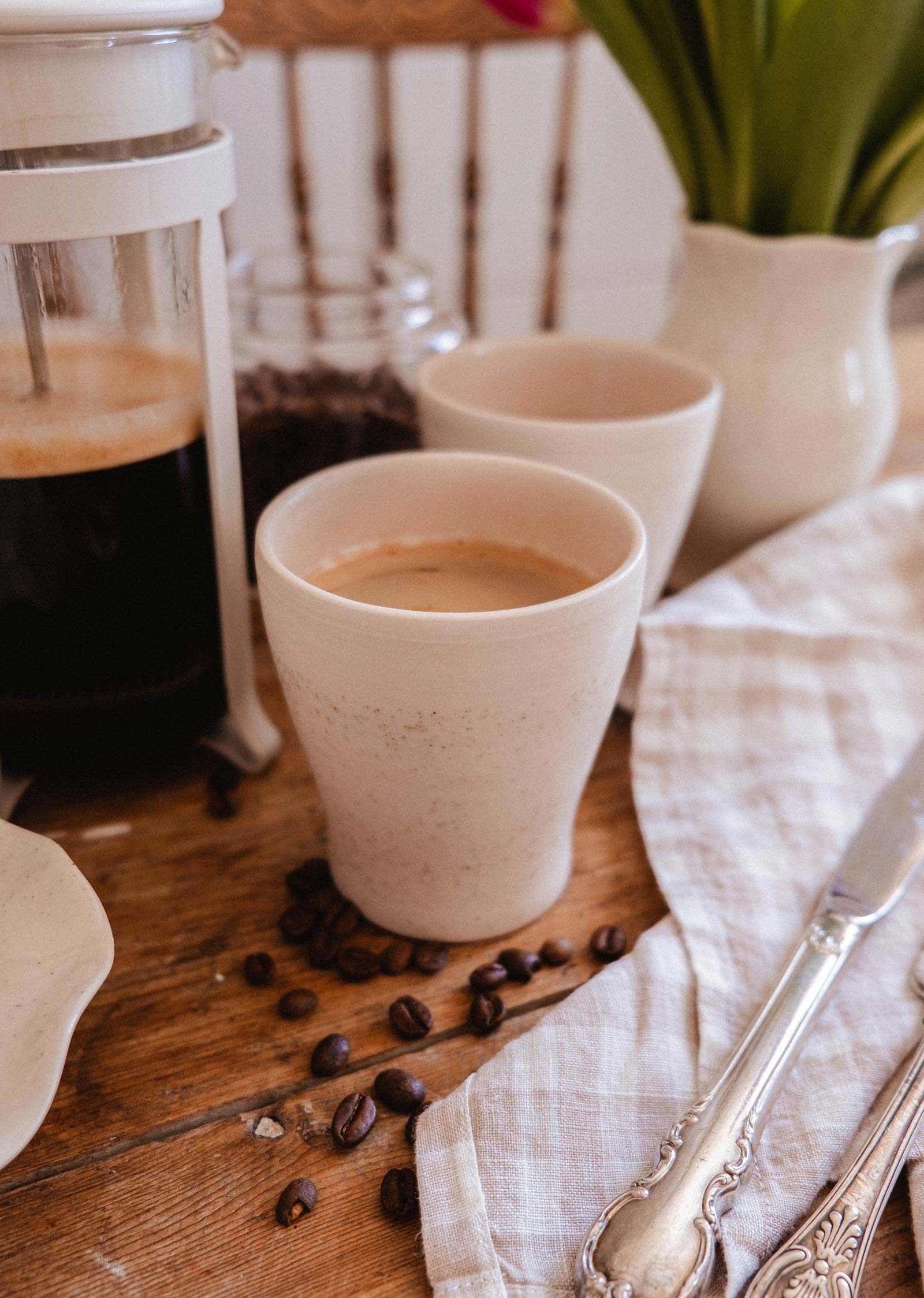 The Small Ceramic Cup by Mimi & August sits on a wooden table with coffee beans, a French press, a napkin, knife, and vase in the background—adding an inviting touch to your home décor.