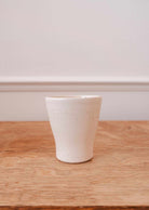 The Small Ceramic Cup by Mimi & August rests on a wooden surface in front of a light-colored wall.