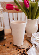 The Tall Ceramic Cup by Mimi & August rests on a wooden table alongside scattered coffee beans, pink tulips in a vase, a French press, and folded napkins with cutlery.