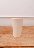 The Tall Ceramic Cup by Mimi & August, handcrafted and off-white, rests on a wooden surface with a neutral backdrop.