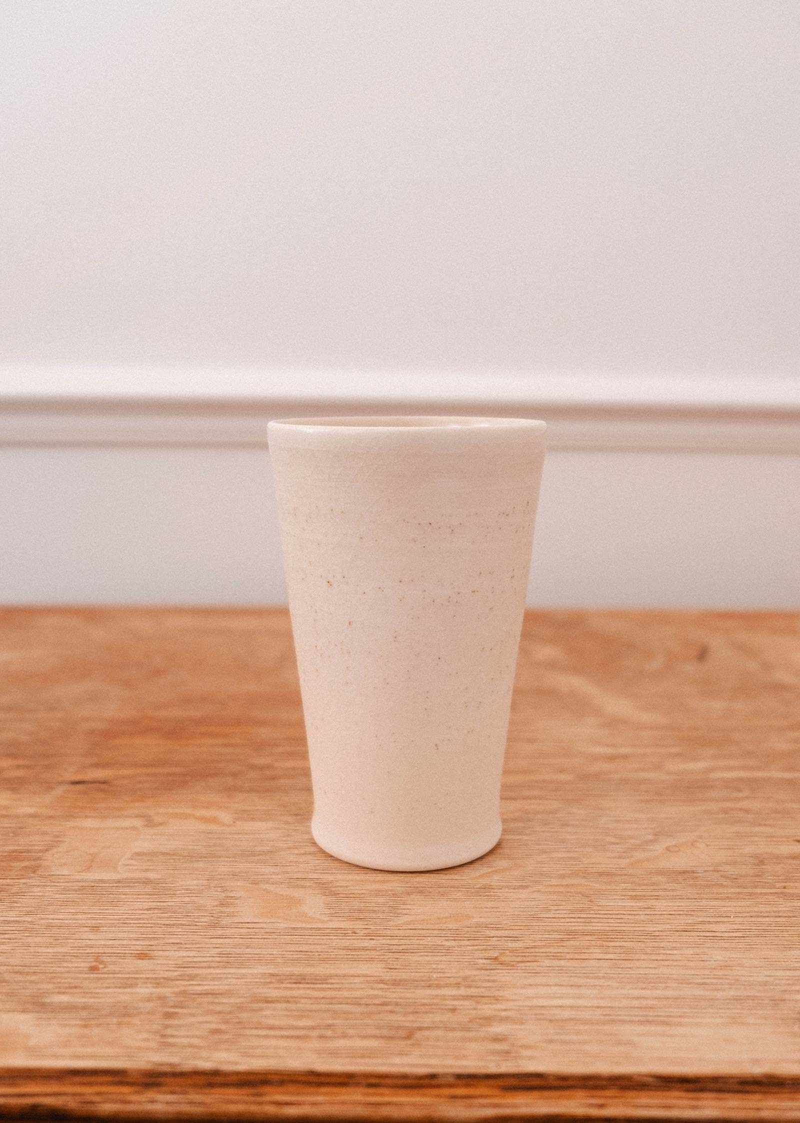 The Tall Ceramic Cup by Mimi & August, handcrafted and off-white, rests on a wooden surface with a neutral backdrop.