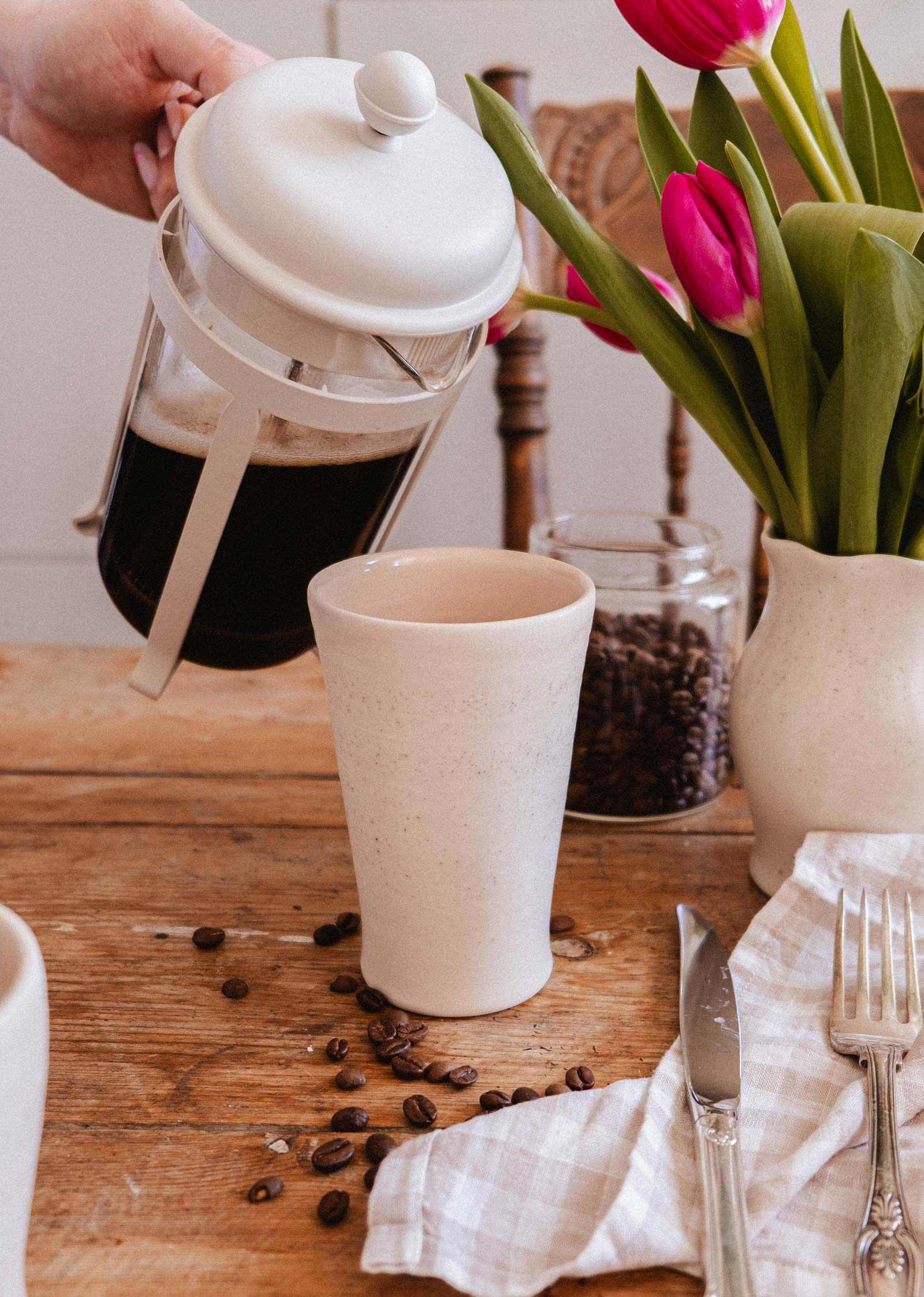 A French press pours coffee into The Tall Ceramic Cup by Mimi & August on a wooden table with coffee beans, cutlery, a checked napkin, pink tulips in a vase, and a jar of coffee beans.