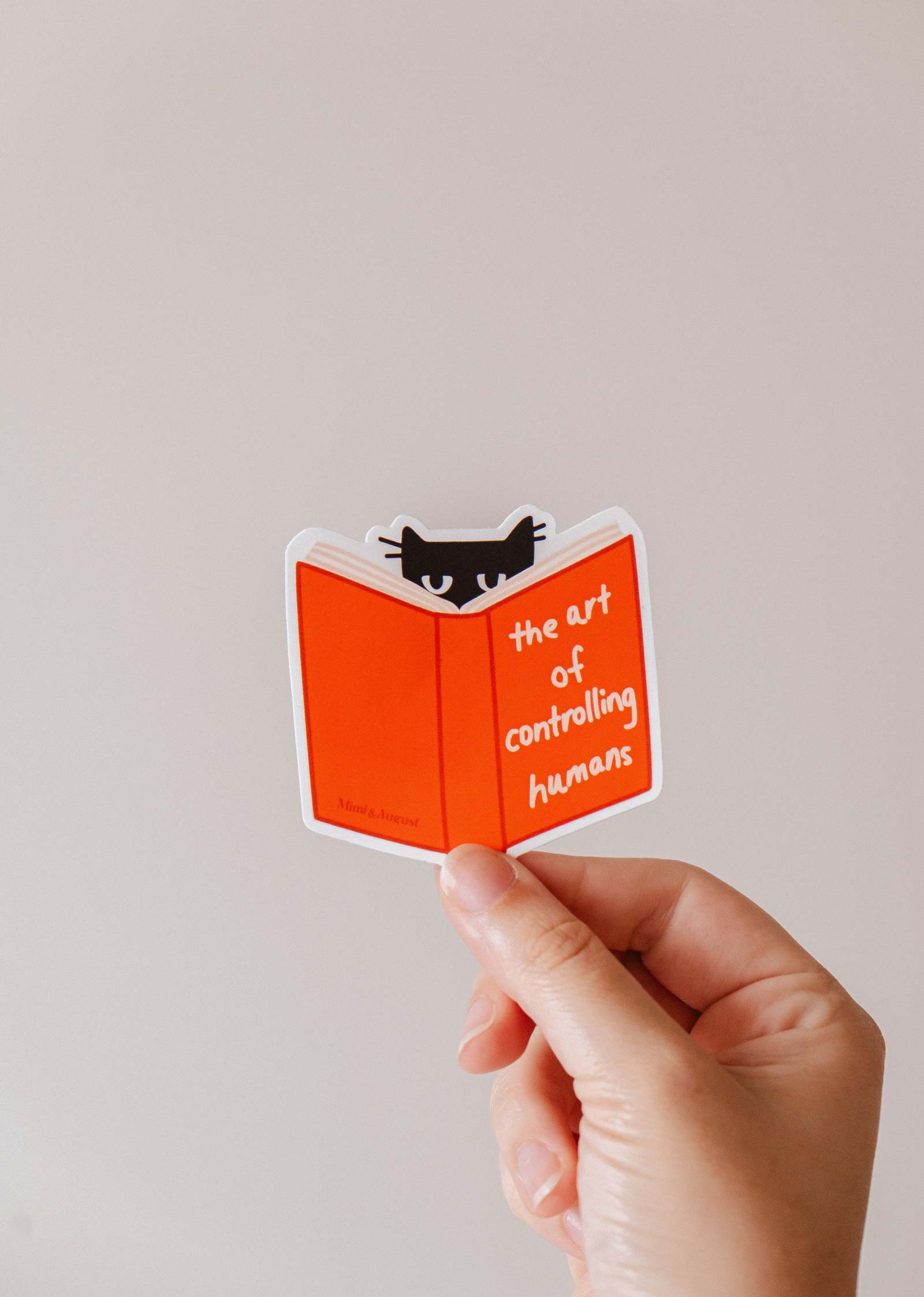 A hand holds Mimi & August's "The art of controlling humans" vinyl sticker, featuring a black cat peeking over an orange book—an ideal quirky gift for cat lovers.