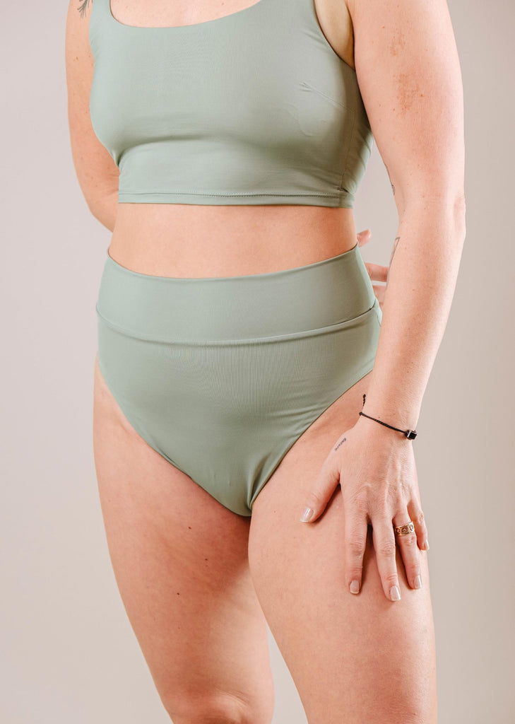 Femme en soutien-gorge de sport vert et bas de bikini Tofino Agave à taille haute de Mimi & August posant avec sa main sur sa hanche sur un fond clair.