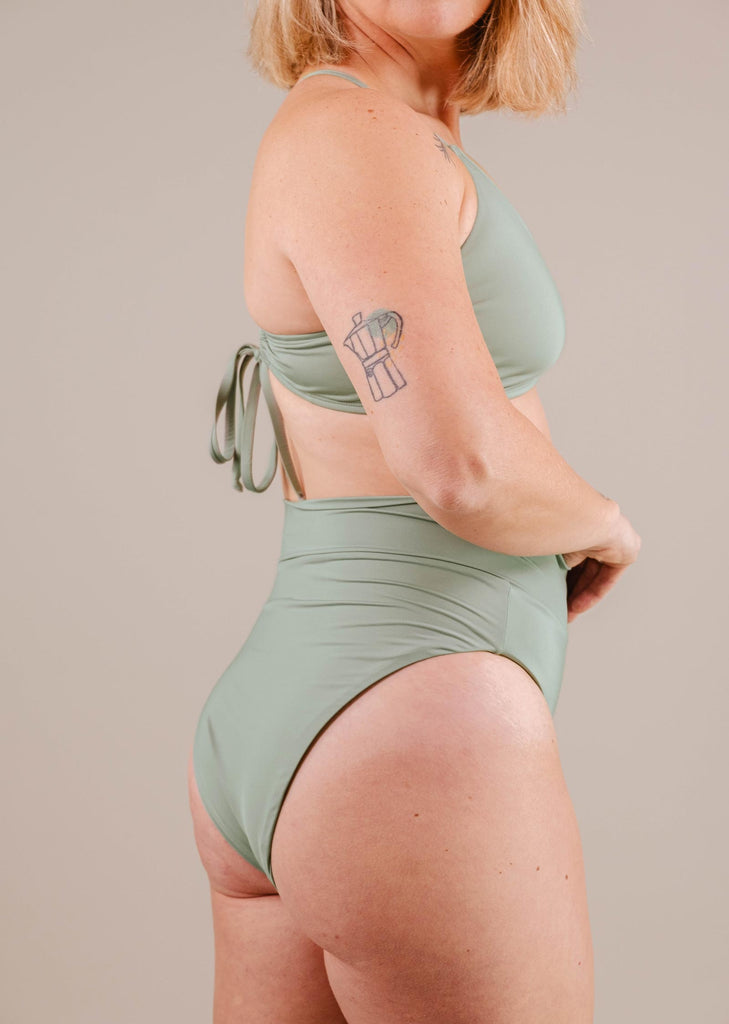 Woman in a Mimi & August Tofino Agave high leg high waist bikini bottom, side pose, showcasing a tattoo on her upper arm.
