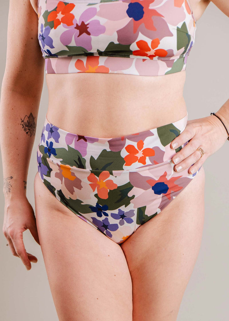 Une personne portant un maillot de bain deux pièces fleuri Mimi & August Tofino Botanica Bas de bikini taille haute cheeky. Gros plan sur le torse et les hanches, mettant en évidence le motif coloré.