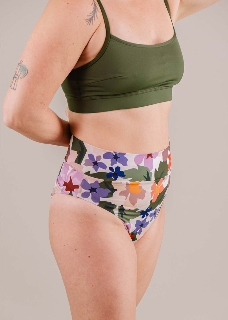 A woman wearing a green sports bra and Mimi & August Tofino Botanica High Leg High Waist Bikini Bottom, viewed from the side against a neutral background. Visible tattoos on her upper arm.