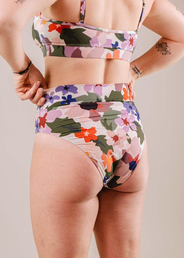 A woman in a colorful, floral Mimi & August Tofino Botanica High Leg High Waist Bikini Bottom posing with hands on her hips against a neutral background.