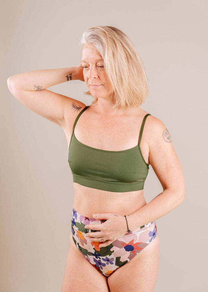 A mature woman in a green sports bra and Mimi & August Tofino Botanica High Leg High Waist Bikini Bottom, posing confidently with her hand on her head, against a neutral background.