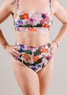 Une femme vêtue d'un soutien-gorge de sport à fleurs et d'un bas de bikini à taille haute Mimi & August Tofino Botanica, debout, les mains sur les hanches, sur un fond neutre. Tatouages visibles sur le haut des bras.