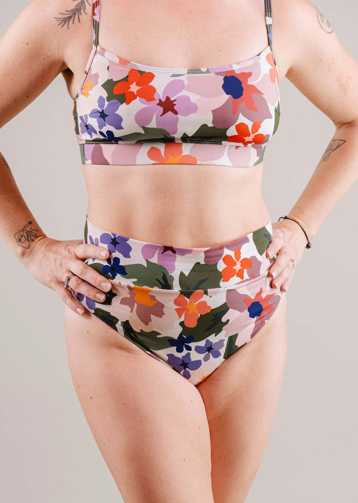 A woman in a floral sports bra and Mimi & August Tofino Botanica High Leg High Waist Bikini Bottom, standing with hands on hips against a neutral background. Visible tattoos on her upper arms.