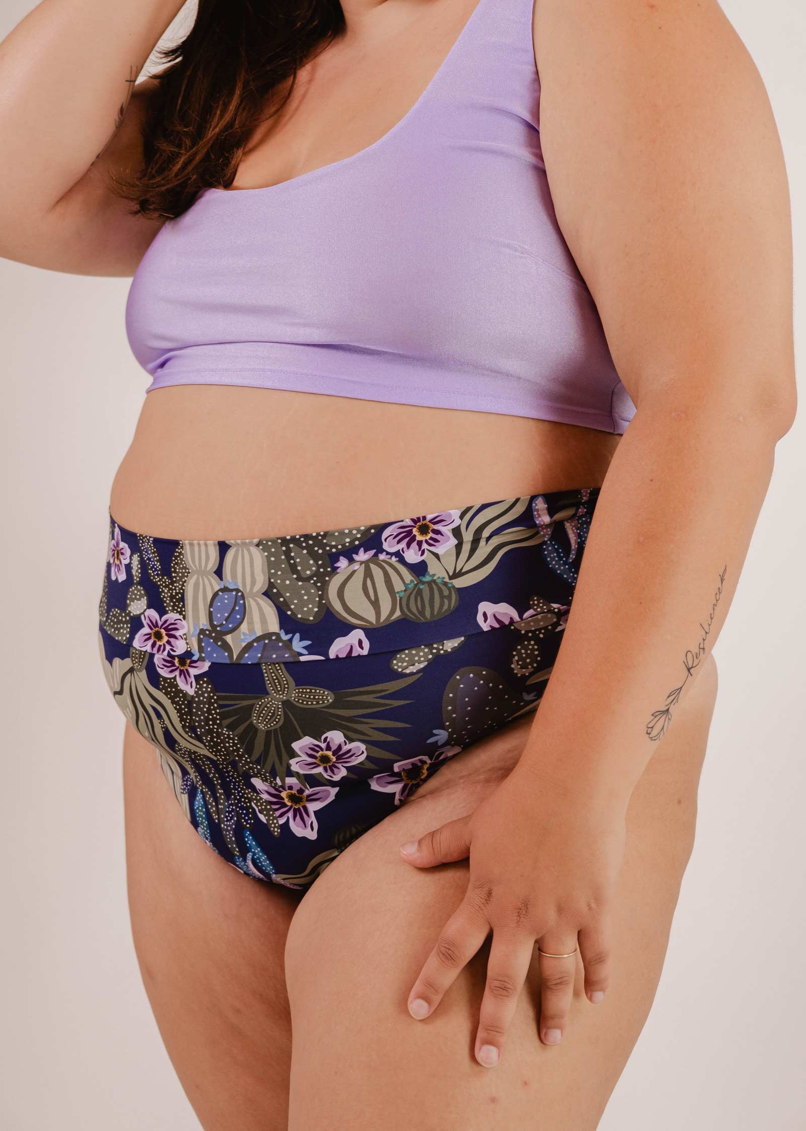 Personne portant un haut de bikini violet et le bas de bikini taille haute Mimi & August Tofino Jardin de Nuit avec des motifs floraux, sur un fond uni.