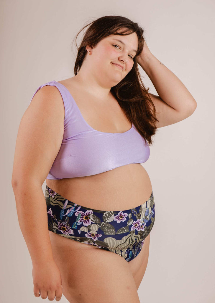 A woman with long dark hair wearing the Mimi & August Tofino Jardin de Nuit High Waist Bikini Bottoms—a purple bikini top paired with floral high-waisted swimsuit bottoms—poses against a plain background.