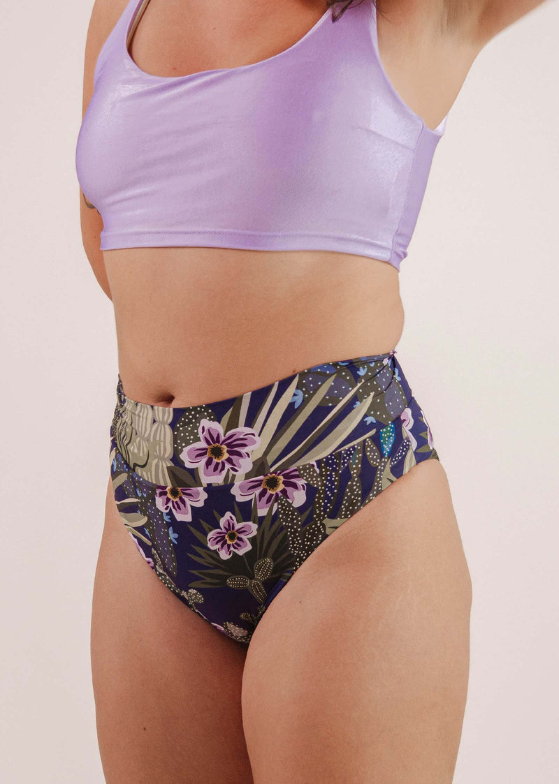 Personne portant un soutien-gorge de sport violet et un bas de bikini taille haute Tofino Jardin de Nuit de Mimi & August sur un fond uni.