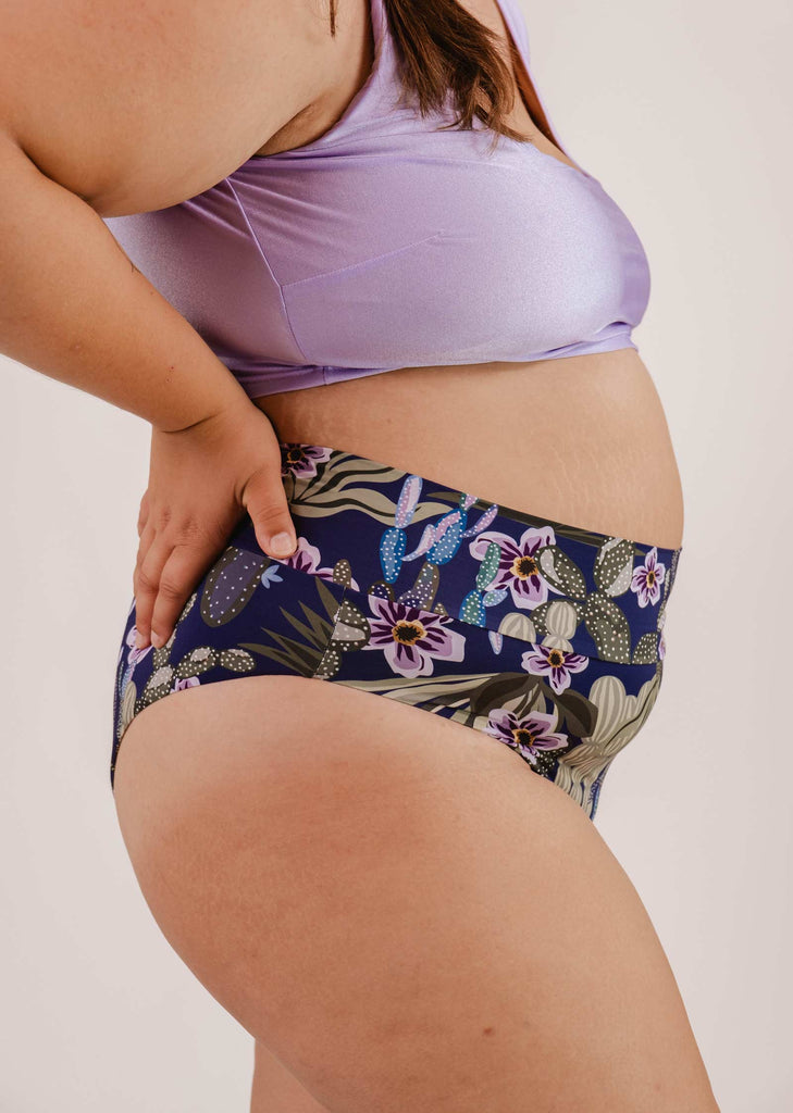 A person wearing a purple crop top and tropical print underwear, akin to the Mimi & August Tofino Jardin de Nuit High Waist Bikini Bottom, with their hand on their hip.