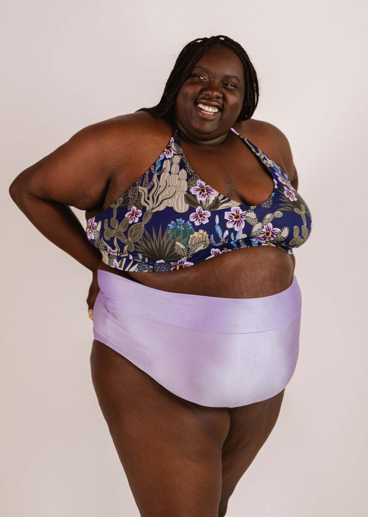 A woman wearing a floral bikini top and Mimi & August Tofino Lilac High Waist Bikini Bottom stands against a plain background, smiling and looking to the side.