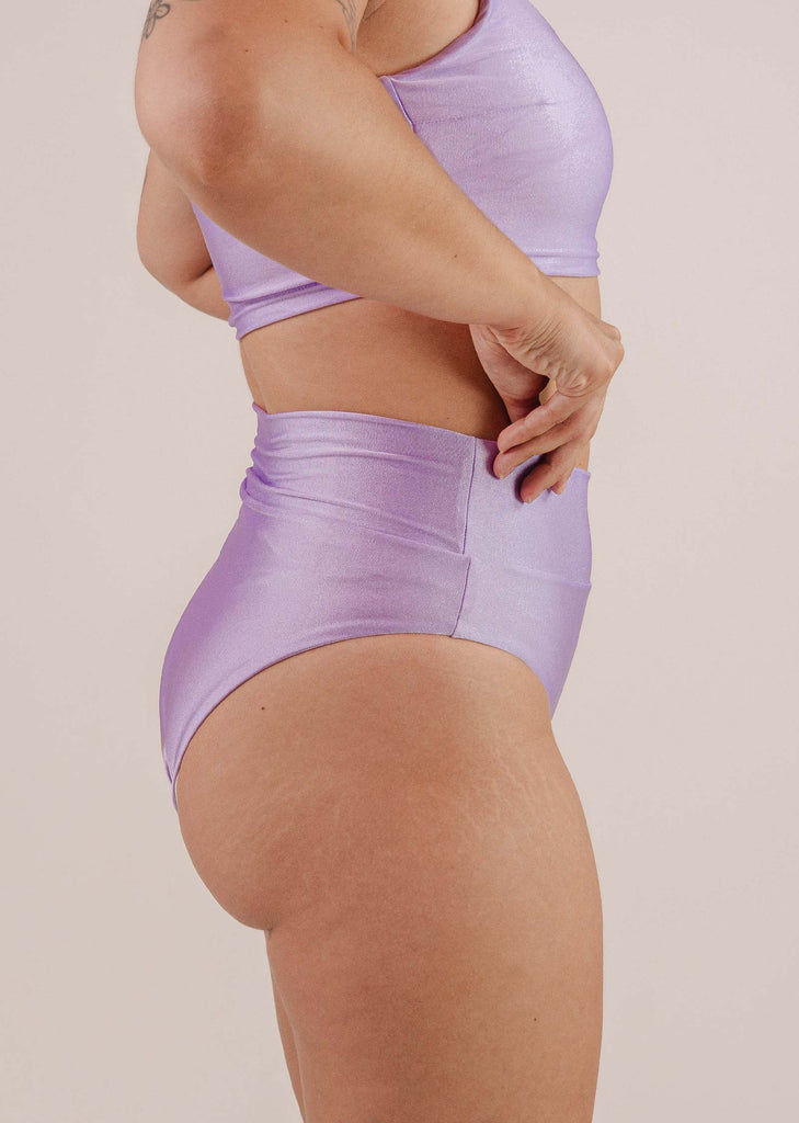 Person standing sideways in a vibrant lilac color, shiny two-piece athletic outfit, with one hand adjusting the waistband of the Tofino Lilac High Waist Bikini Bottom by Mimi & August.