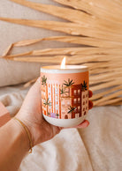 A hand holds a lit Topaze - Reusable Candle by Mimi & August in a white container with an orange and pink design featuring buildings and leaves, creating a tranquil ambience.