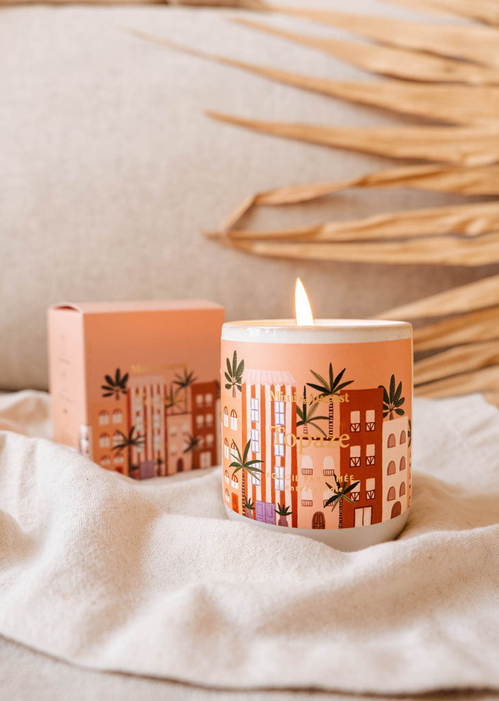 A lit Topaze - Reusable Candle from Mimi & August, featuring a cityscape design, sits on a fabric surface exuding a sophisticated sandalwood scent. A matching box is visible in the background, with dry palm fronds partially in view behind the candle.