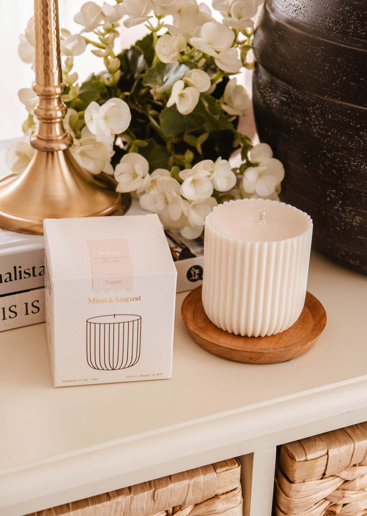 A ribbed, white candle in a glass sits on a wooden coaster beside its box, which reveals it as the Candle Refill - Topaze by Mimi & August. It’s surrounded by decorative items including a brass lamp and white flowers, enhancing the serene ambiance with hints of sandalwood coconut vanilla.