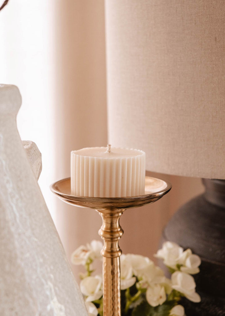 A white, ribbed pillar candle sits on a gold holder beside a white ceramic vase and a gray lamp, with white flowers partially visible in the background. The candle is actually a Candle Refill - Topaze by Mimi & August with a delightful sandalwood coconut vanilla fragrance, highlighting an eco-friendly choice.