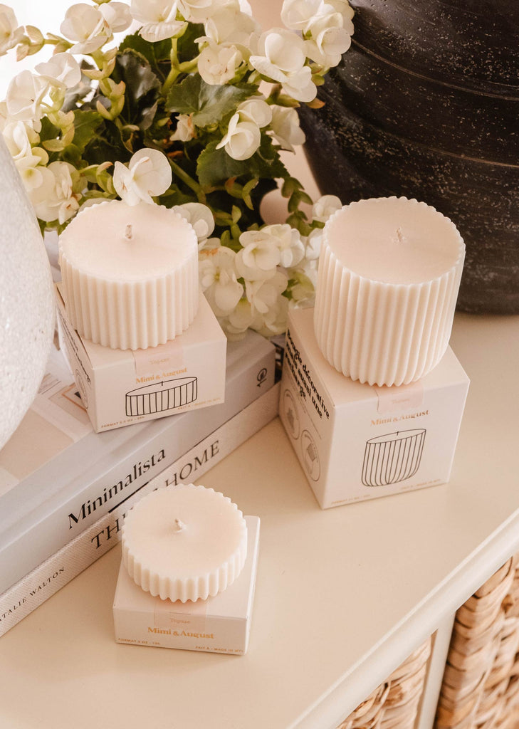 Three decorative candles in ribbed white holders are arranged on boxes and stacked books next to a vase of white flowers, exuding the delightful scents of sandalwood, coconut, and vanilla. Consider eco-friendly candle refills like Candle Refill - Topaze from Mimi & August for a sustainable touch.