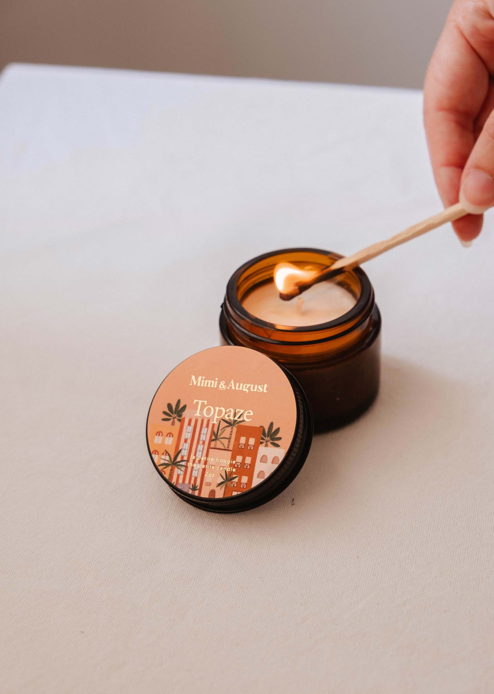 A hand lights the Mimi & August Topaze - 2 oz coco vanilla candle in a brown glass jar, with its illustrated reusable lid nearby on a light surface.