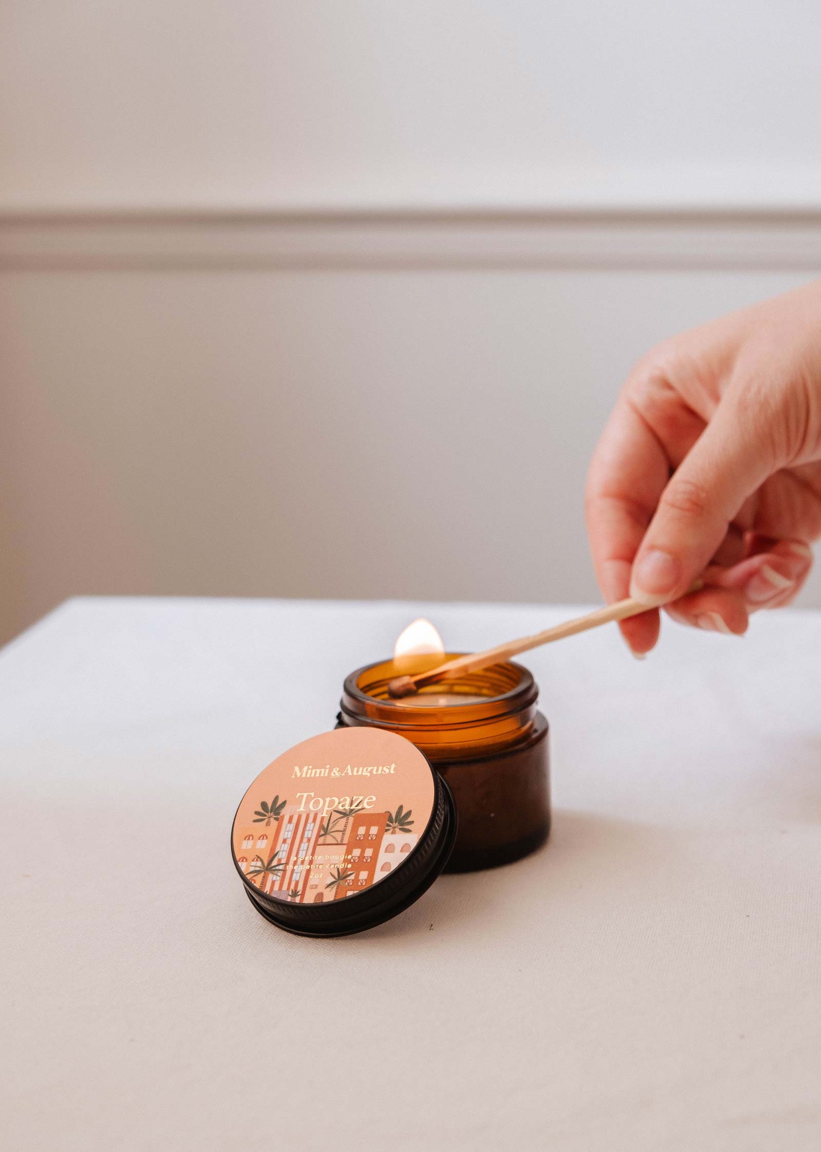 A hand lights Mimi & August's Topaze - 2 oz sandalwood candle in a brown glass jar on a white surface, with the lid nearby.
