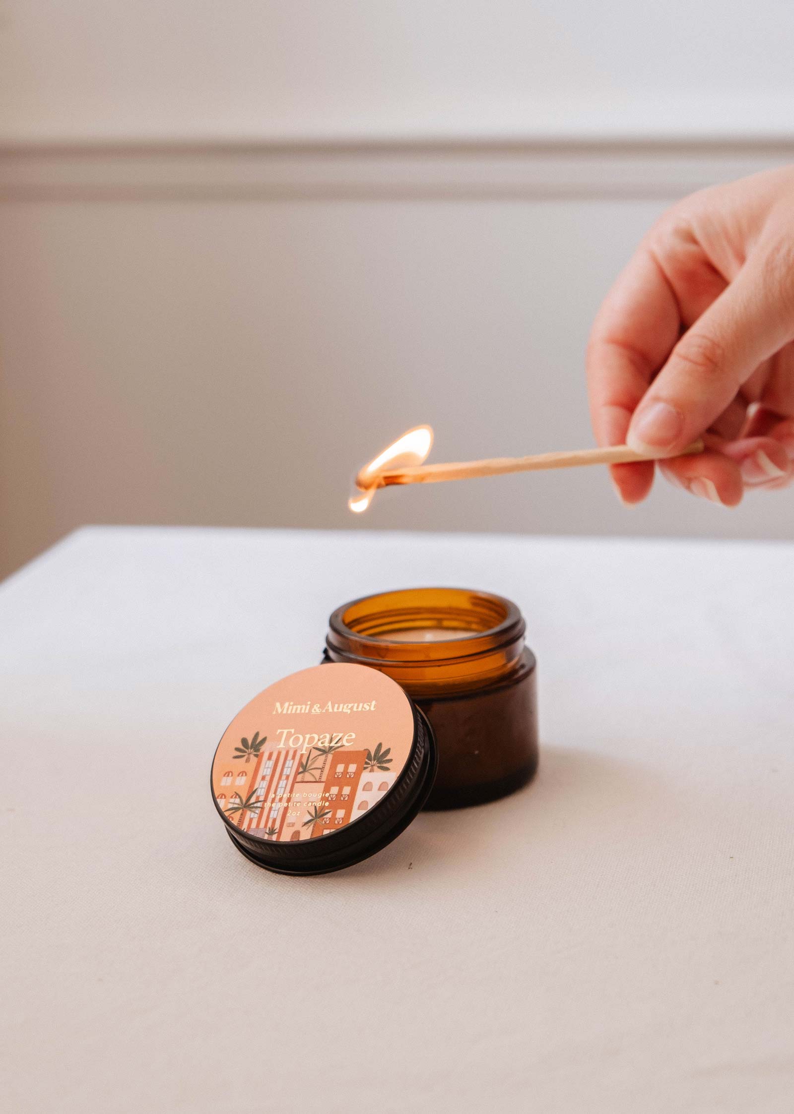 A hand lights a wooden match above an open amber jar candle labeled "Topaze - 2 oz" by Mimi & August on a white surface, filling the air with soothing scents—a tranquil oasis in a reusable candle.