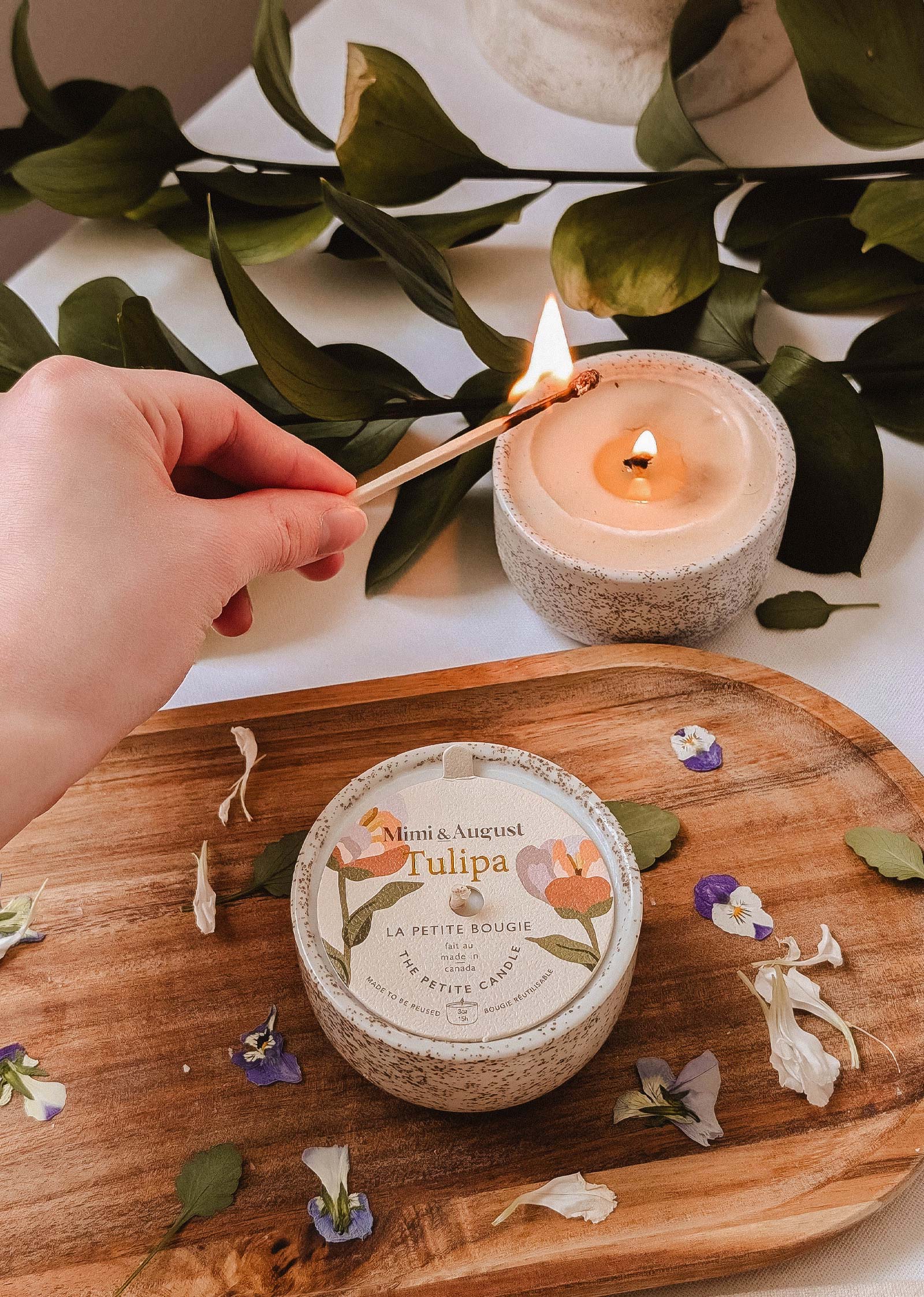 A hand lights the Mimi & August Tulipa - Reusable Candle with wildberries fragrance on a wooden tray next to another candle and scattered flower petals, with green leaves in the background.