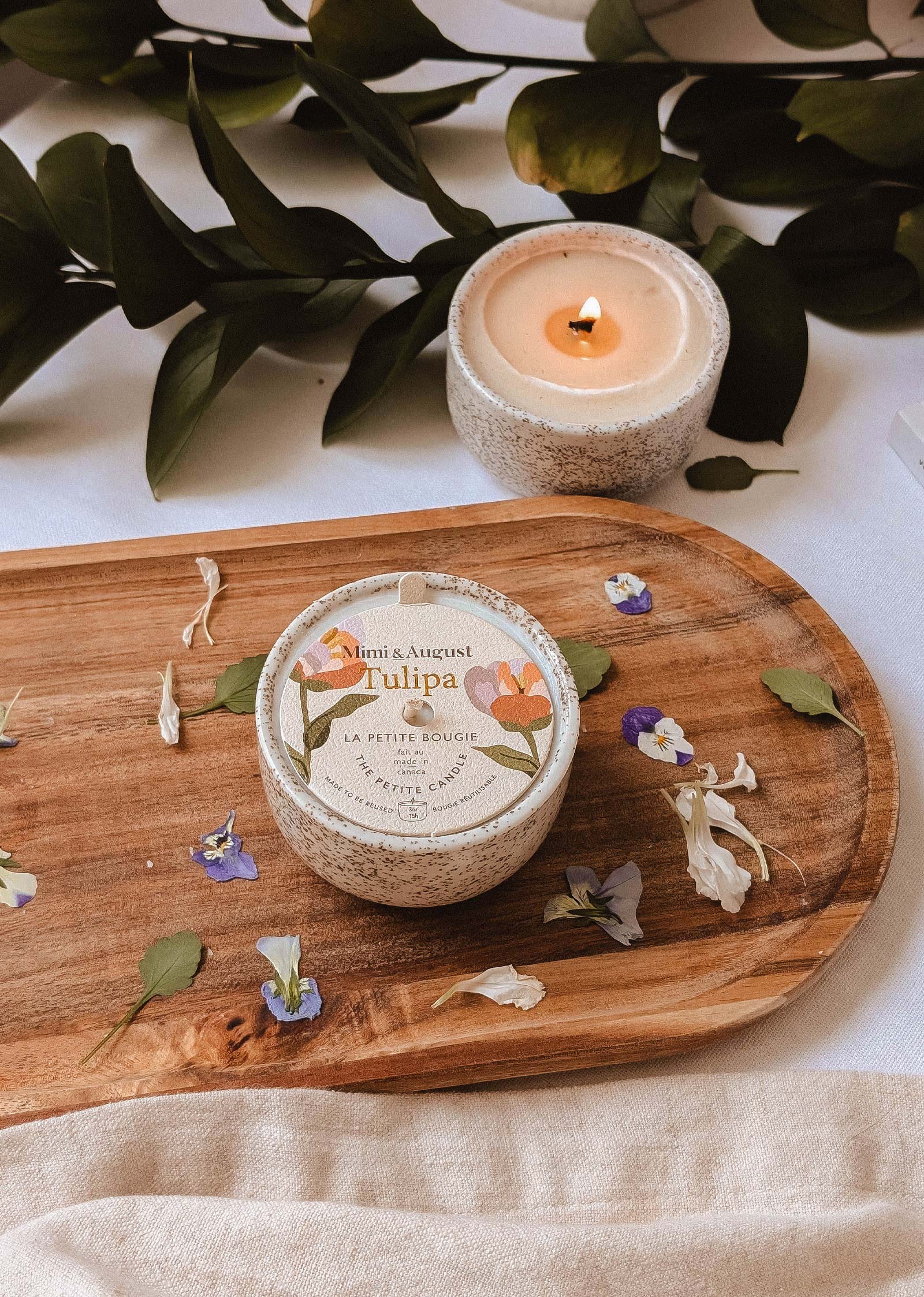 The Tulipa - Reusable Candle by Mimi & August sits round on a wooden board amid scattered petals, with a second lit reusable candle nearby. Greenery in the background enhances the charming scene.