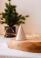 A small ceramic tree, akin to the Twinkle Tealight Holder by Mimi & August with its star-shaped cutouts, is perched on a wooden platform. In the softly lit background, a potted evergreen wrapped in string lights sparkles gently, enhancing the festive Christmas decor.