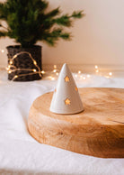 The Twinkle Tealight Holder by Mimi & August, a small ceramic cone with star-shaped cutouts, is illuminated from within and positioned on a wooden surface. In the background, a softly blurred potted plant and string lights add to the cozy Christmas decor ambiance.