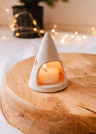 The Twinkle Tealight Holder from Mimi & August is a charming ceramic candle holder designed in a cone shape with star-shaped cutouts, ideal for holding a glowing tealight candle. Set elegantly on a wooden surface with softly blurred fairy lights twinkling in the background, it makes for exquisite Christmas decor.