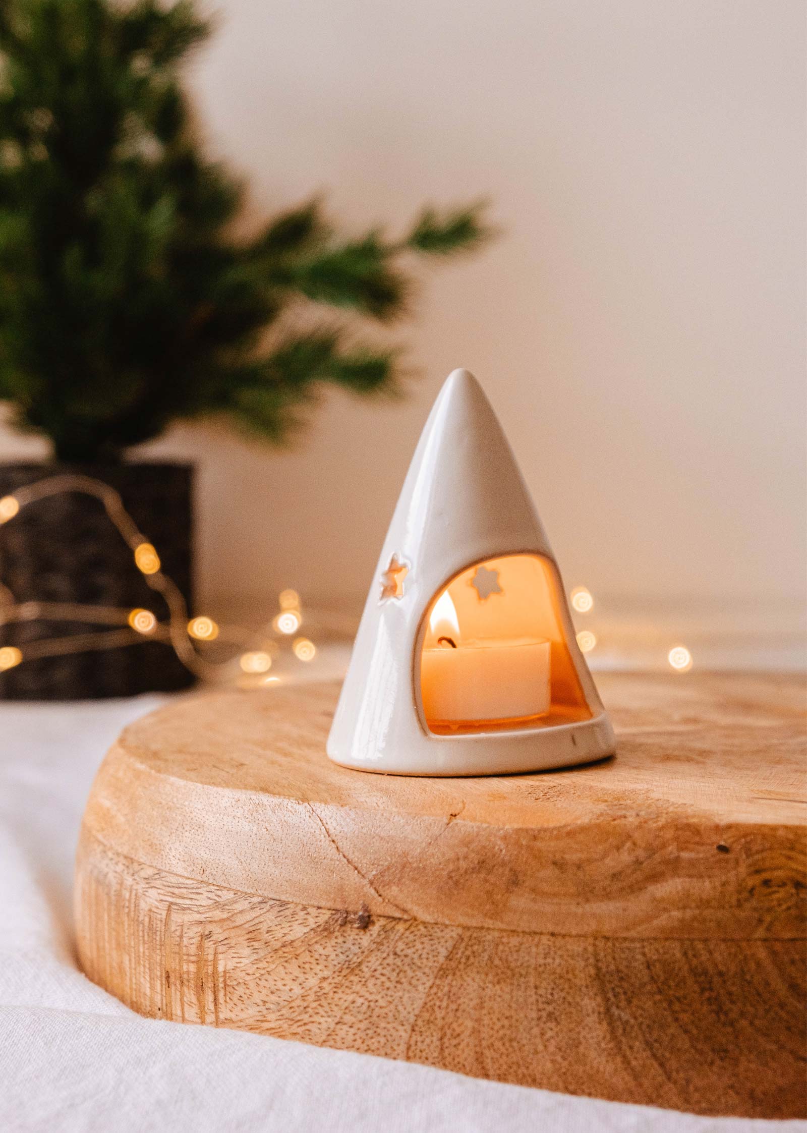 Un petit porte-bougies à réchaud Twinkle de Mimi & August, de forme conique, avec des découpes en forme d'étoile, est posé sur une surface en bois avec une bougie allumée à l'intérieur. Un arbre flou et des guirlandes lumineuses en arrière-plan renforcent l'ambiance de la décoration de Noël. Idéal pour ajouter un charme festif.