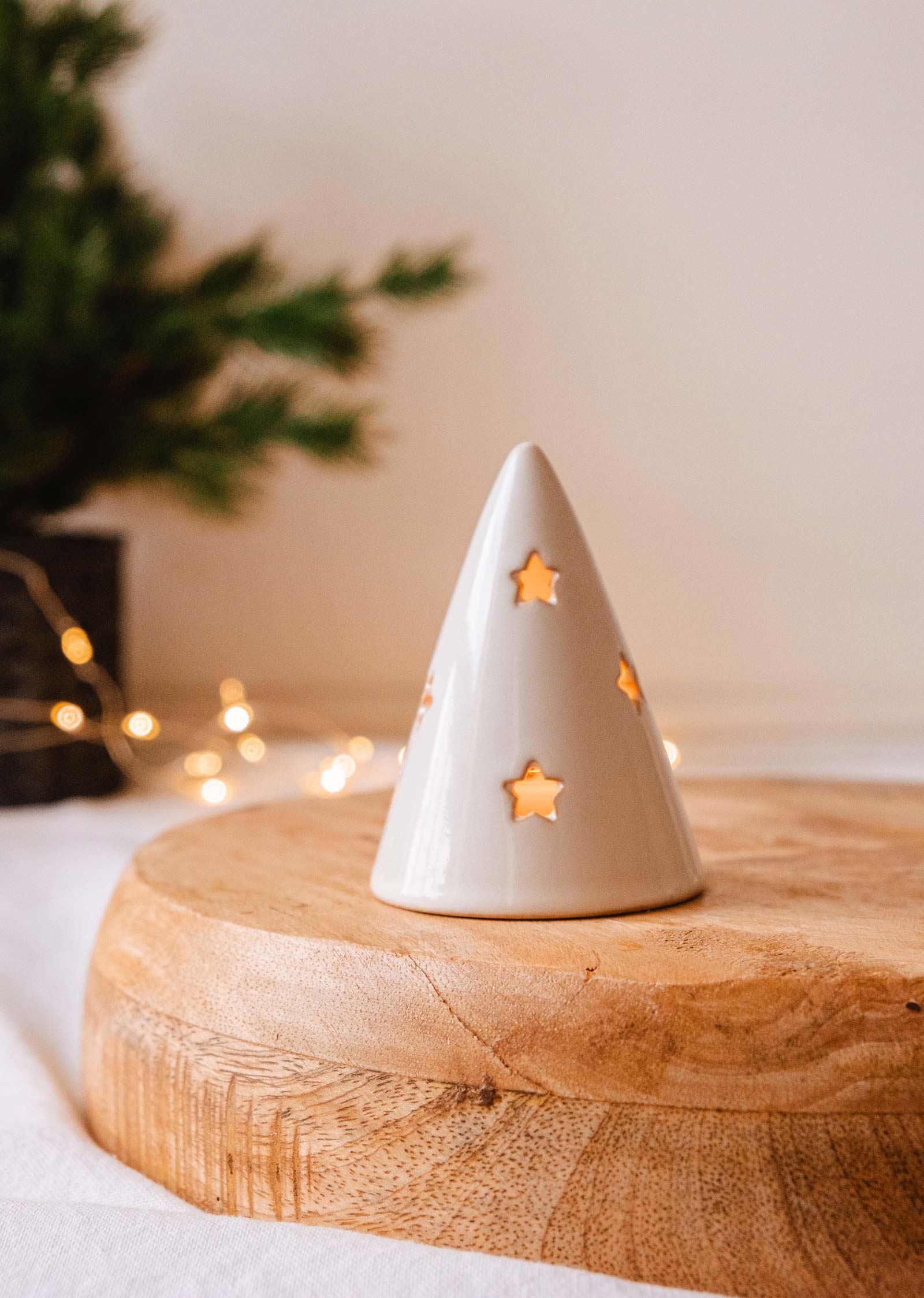 The Twinkle Tealight Holder by Mimi & August, featuring star-shaped cutouts, rests elegantly on a wooden surface. Its cozy glow illuminates against blurred fairy lights and vibrant greenery, bringing a touch of Christmas decor magic to any setting with this delightful ceramic tealight holder.