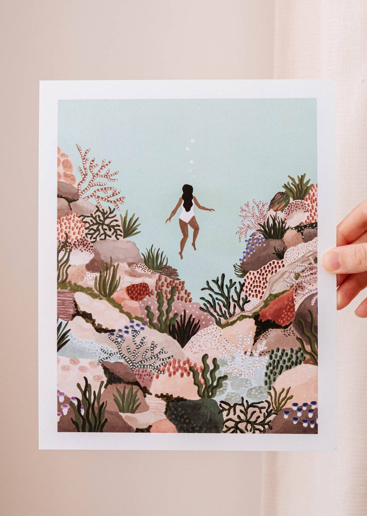 A woman holding up a print of a woman swimming in the underwater world, featuring the Under the sea Art Print by Mimi & August.