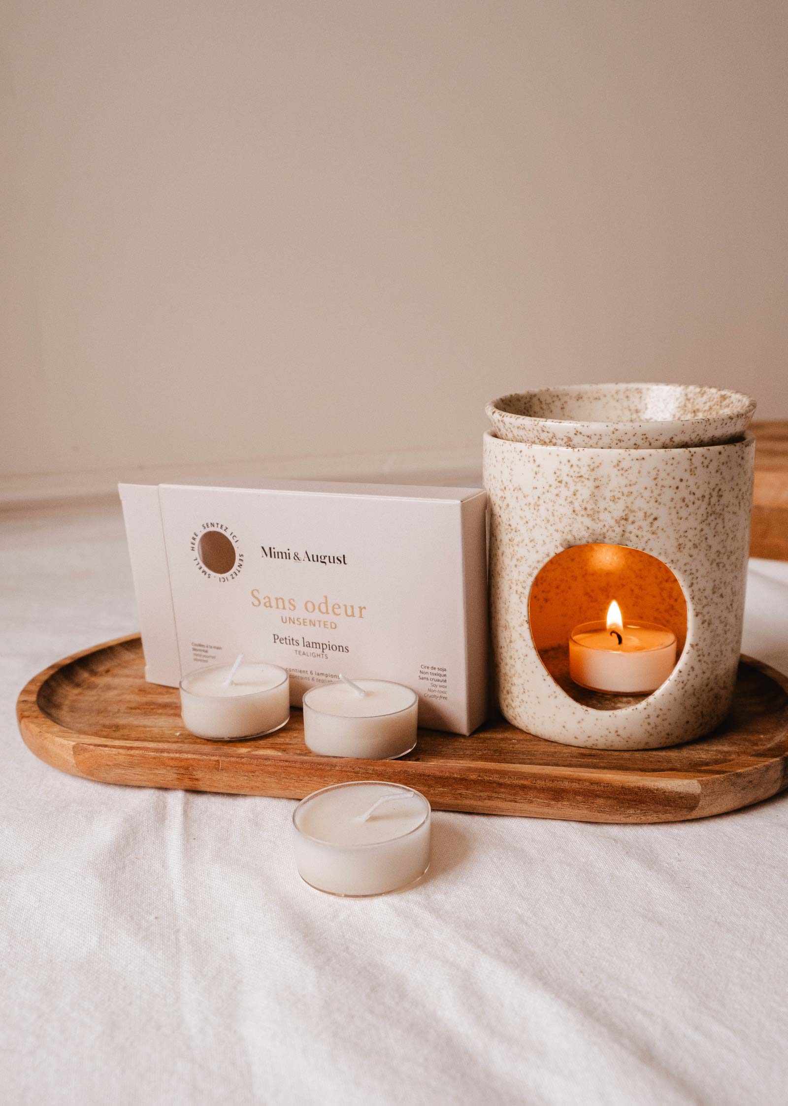 A ceramic holder with a burning tealight from the Unscented Tealight Candle Set by Mimi & August sits on a wooden tray beside a "Sans odeur" box and more candles, providing clean, steady light for any space.