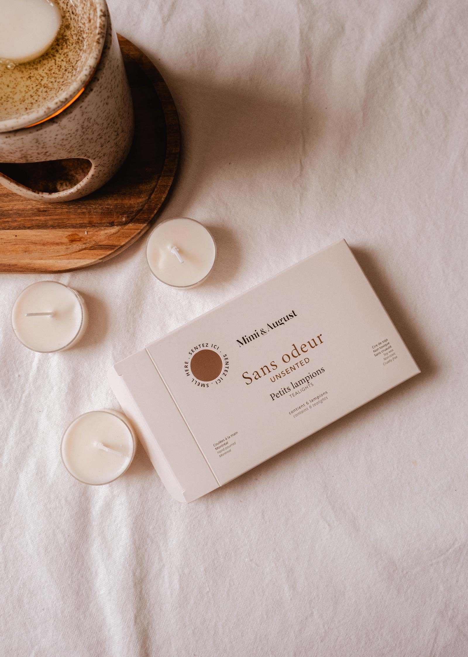 A Mimi & August Unscented Tealight Candle Set with pure soy wax tealights and a ceramic mug on a wooden coaster, arranged on a white fabric surface.