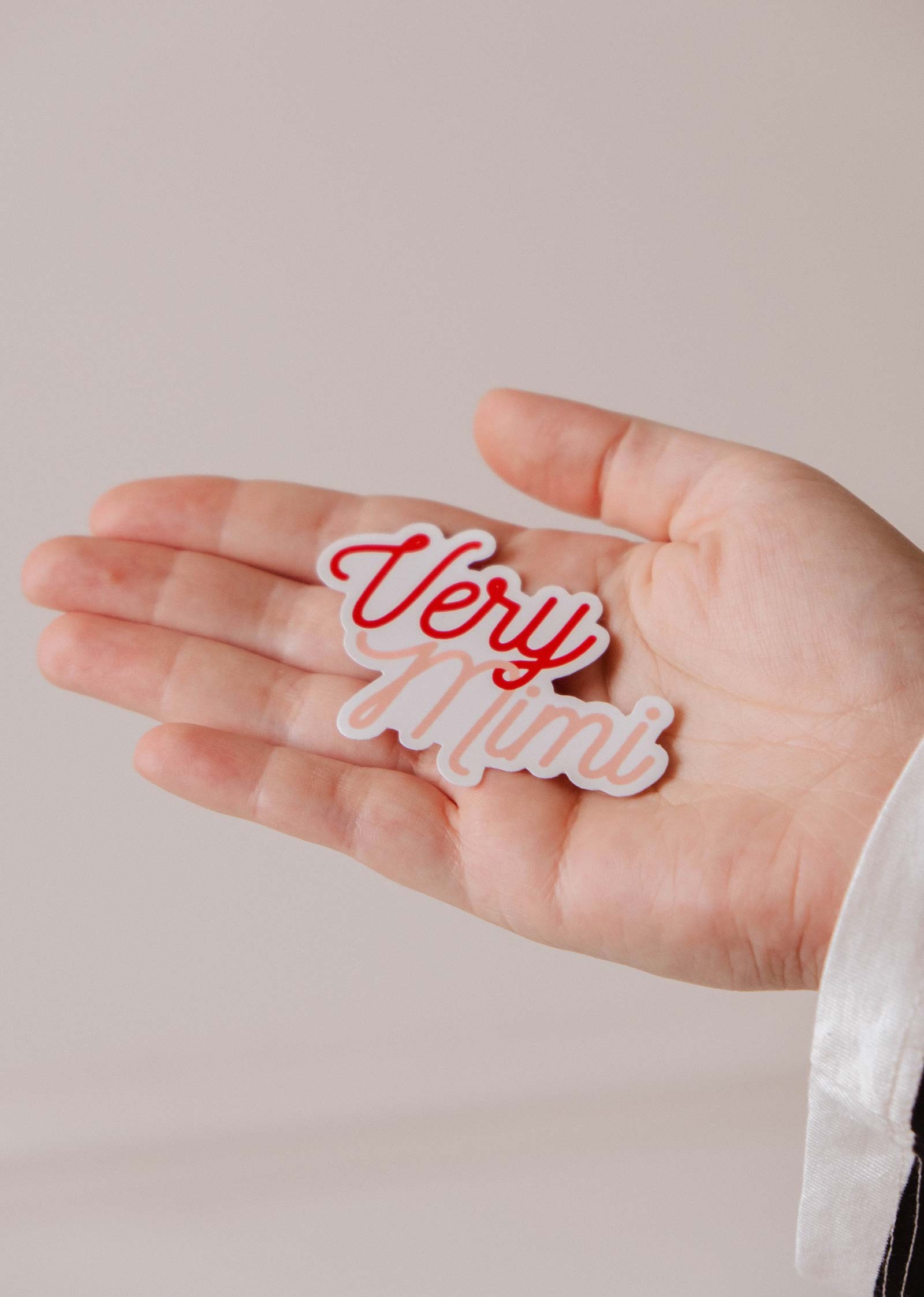 A hand holds a "Very Mini" cutout in red and white, reflecting Mimi & August’s playful style—a perfect match for the Very Mimi - Vinyl Sticker by Mimi & August, designed by Camille Forcherio.