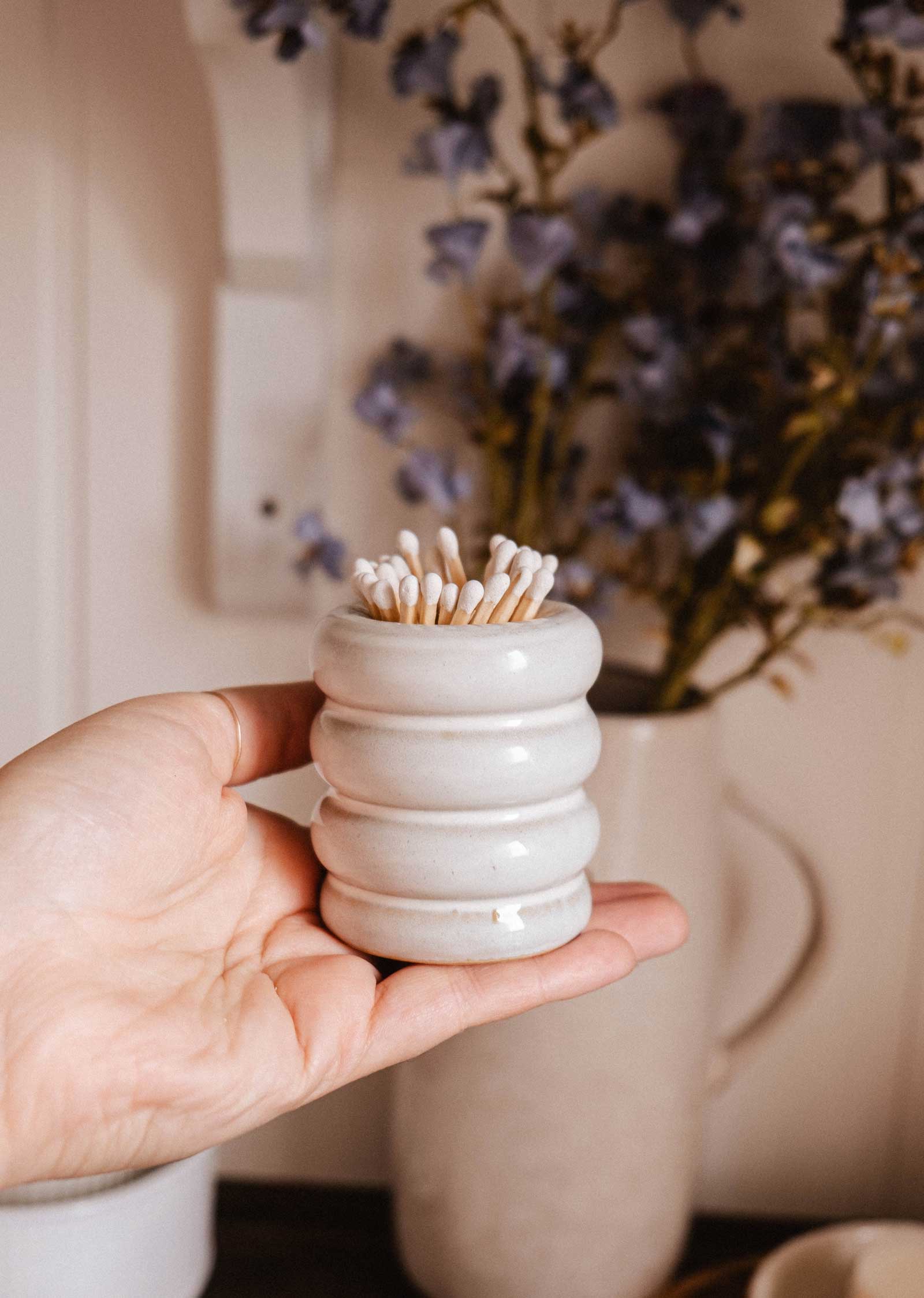 A hand holds the Mimi & August Ceramic Match Pot filled with cotton swabs; blurred purple flowers in a vase appear in the background.