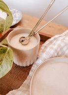 The Mimi & August Wick Trimmer neatly trims candle wicks, shown here with a white candle in a beige ceramic container on a wooden surface—an essential accessory for any cozy space.