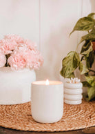 A Mimi & August Special Edition Candle glows on a woven mat next to a vase of pink flowers, a green potted plant, and a small cotton swab container.