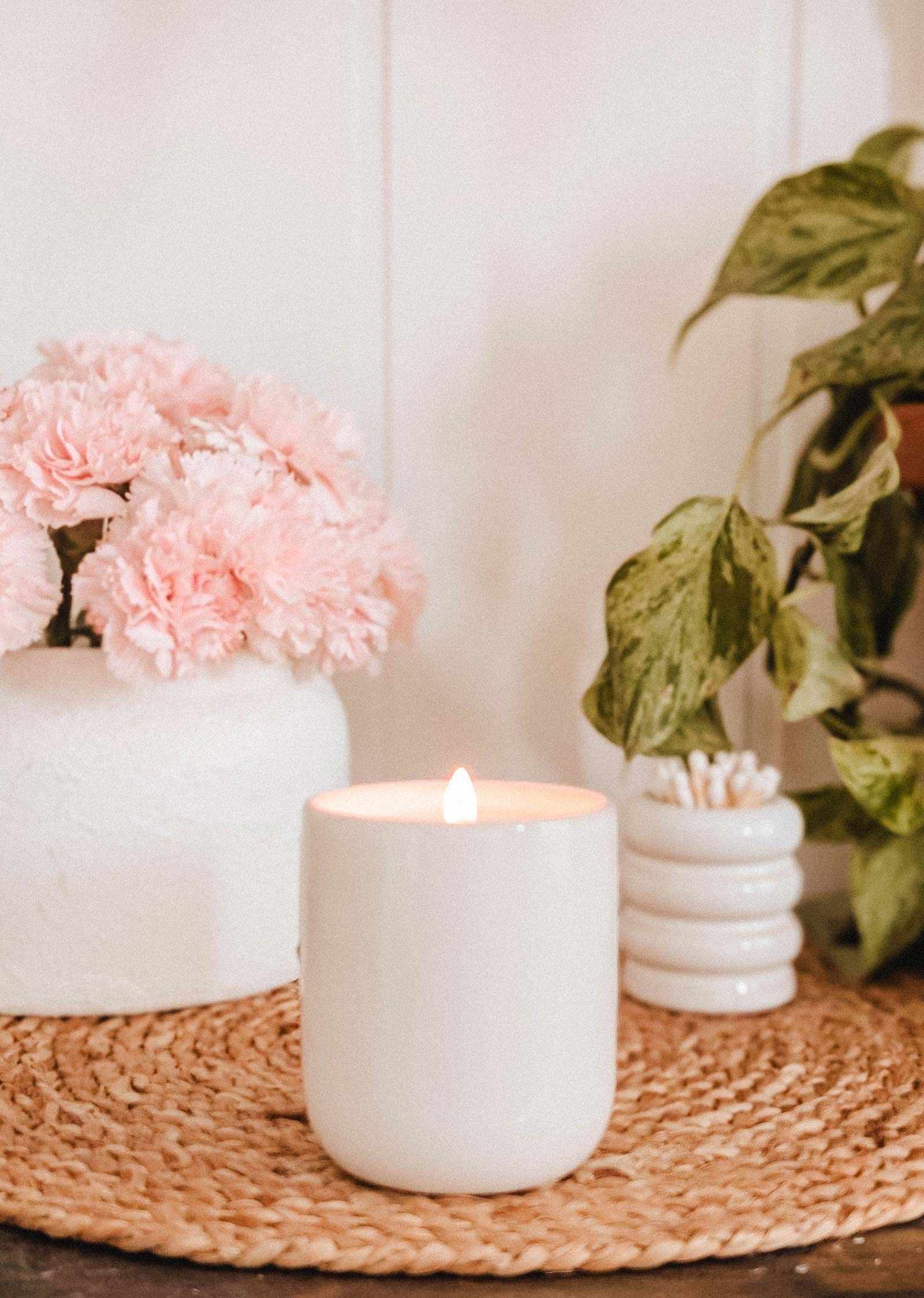 A Mimi & August Special Edition Candle glows on a woven mat next to a vase of pink flowers, a green potted plant, and a small cotton swab container.