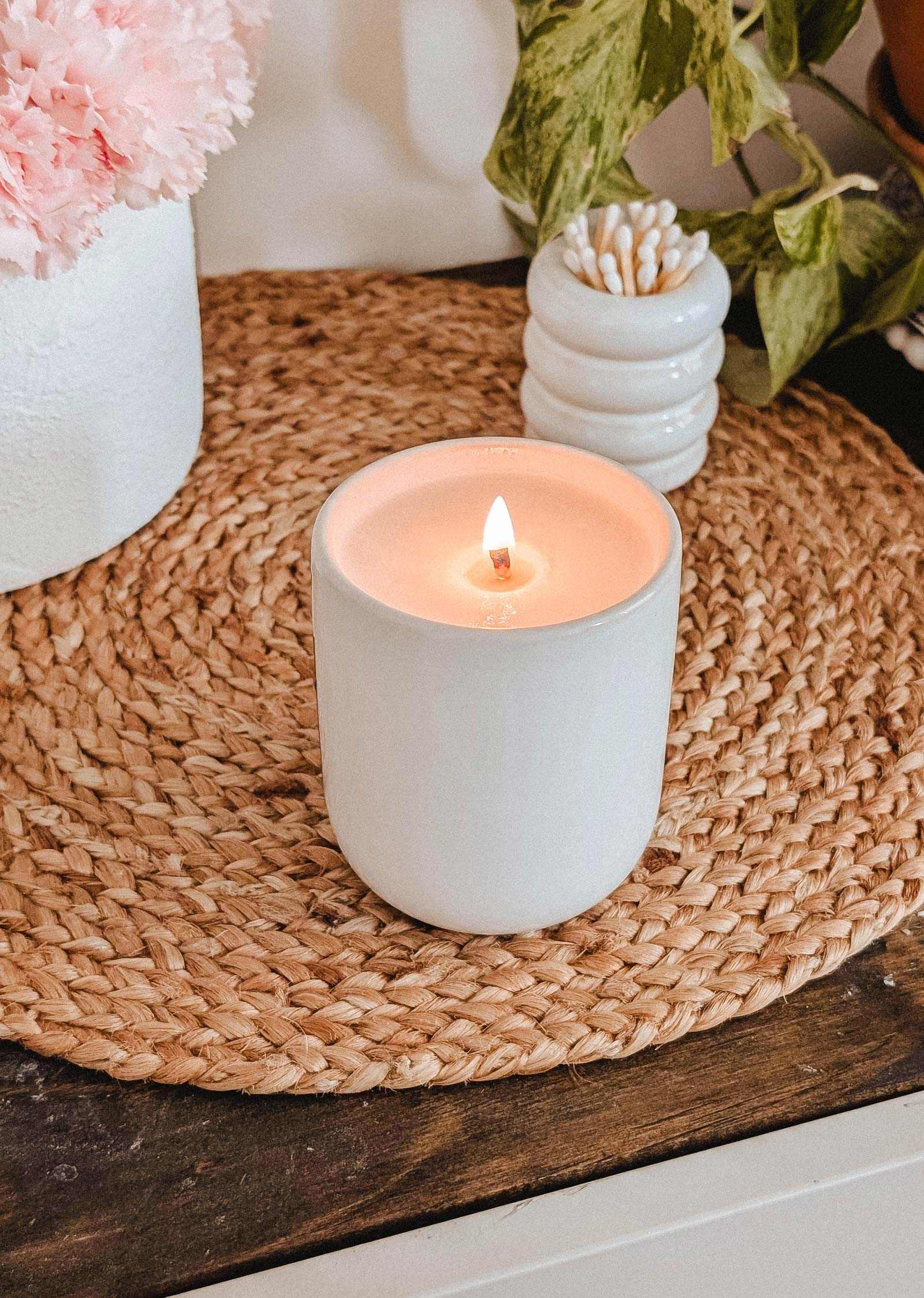 The Special Edition Candle by Mimi & August glows in a ceramic jar on a woven placemat, beside a white vase filled with pink flowers and a small cotton swab container.