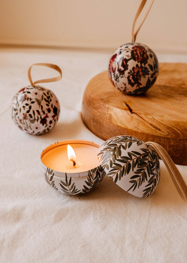 A *Wintea - Christmas Ornament Candle* by Mimi & August, featuring a leafy tin lid, sits on a tablecloth. In the background, two decorative spheres with floral patterns mimic elegant Christmas baubles, with one placed on a wooden board.