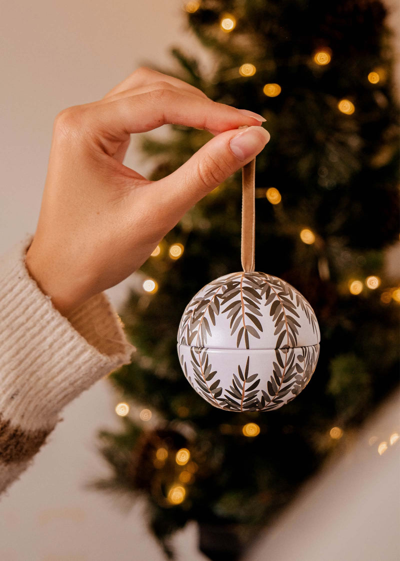 A hand holds the Wintea - Christmas Ornament Candle by Mimi & August, featuring leaf patterns reminiscent of festive baubles, in front of a lit Christmas tree.