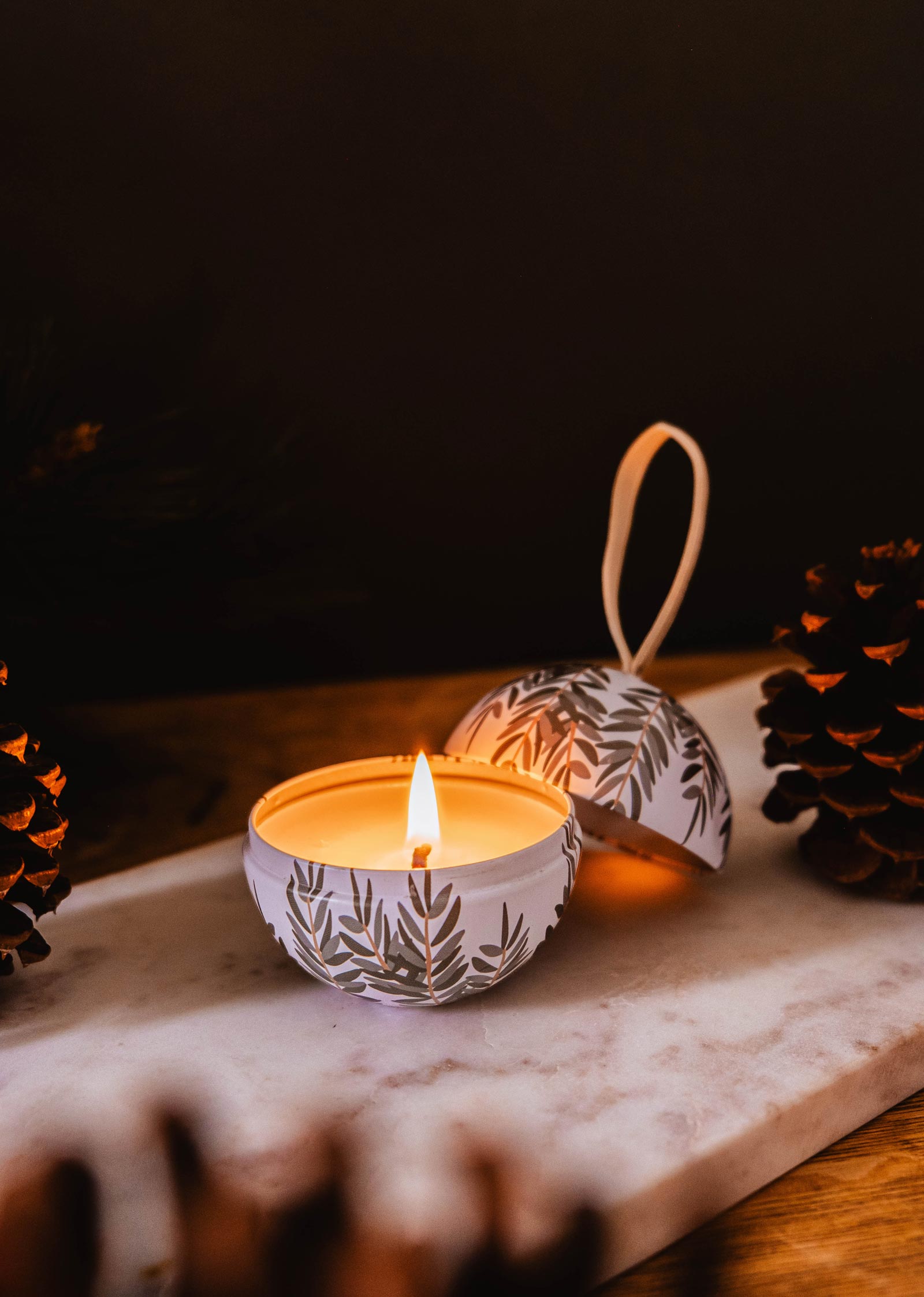 A Wintea - Christmas Ornament Candle by Mimi & August, elegantly housed in a decorative tin adorned with leaf patterns, rests on a marble surface amidst pinecones and delicate holiday baubles.